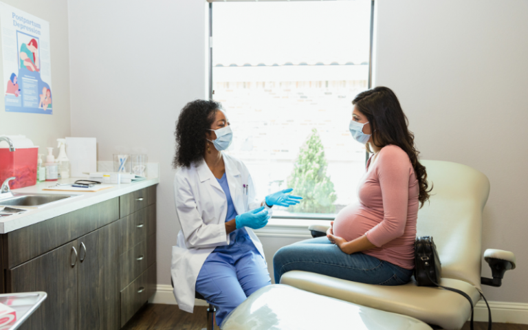 At the prenatal appointment during the coronavirus epidemic, the mature adult female doctor with the protective mask gestures and talks to her pregnant patient.