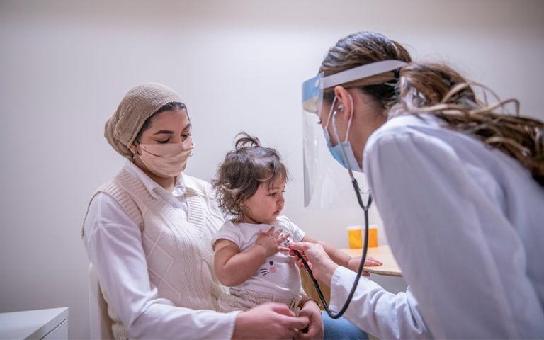 A masked female doctor is using a stethoscope to listen to a toddler girl's chest. The young patient is sitting on her Muslim mother's lap at the medical office.