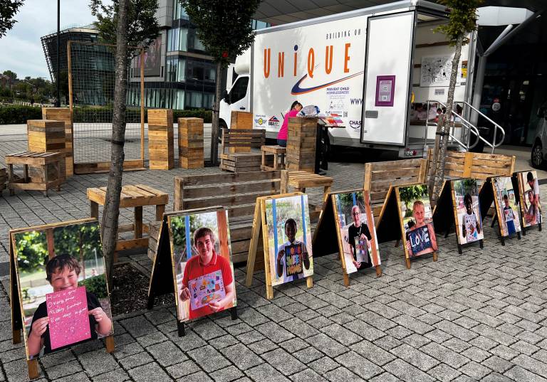 Public square with a van parked and outside the van are displayed a series of sandwich boards with images and message from children fixed on the boards