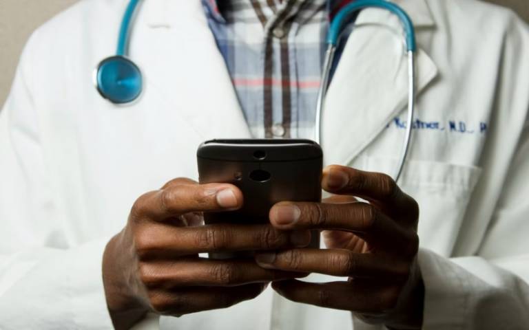 A doctor with a stethoscope and white scrubs on holds a phone - only hands and torso shown