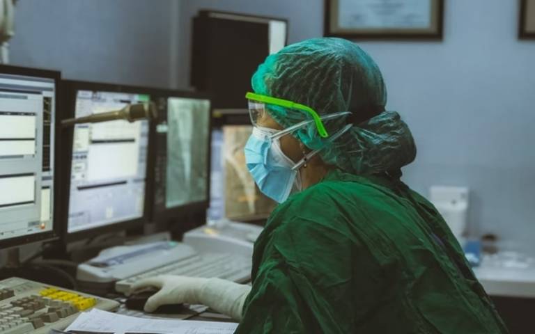 Hospital researcher in green scrubs looks at three screens of medical data