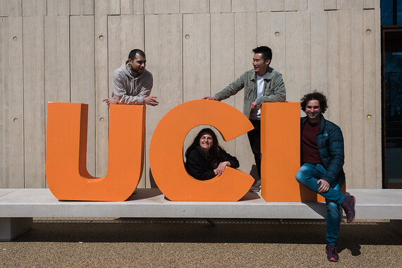 Students leaning on 3d letters in orange spelling UCL on a grey concrete background