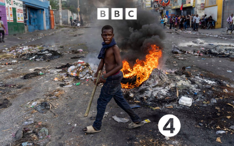 a black child walks amongst rubble with a fire behind him