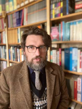 a man with a beard, glasses wearing a blazer stands in front of a bookcase