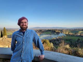 a white man with a beard is wearing a blue striped shirt and pink beret. There is a beautiful countryside view behind him.