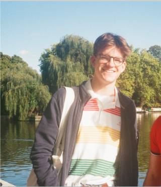 A young man with brown hair and glasses smiles at the camera with trees and a body of water behind him