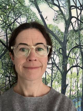 A white middle aged woman in clear glasses and a grey jumper her brown hair tied back smiles at the camera against a backdrop of painted trees.