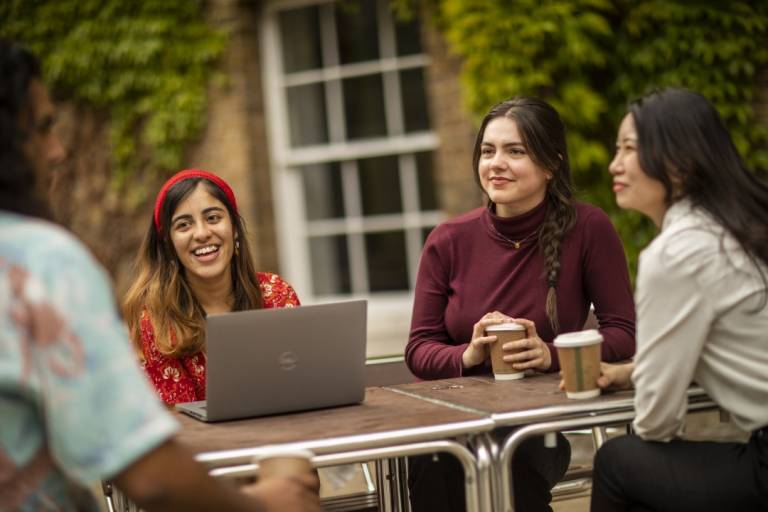 A group of four students chat and have coffee around an outdoor table. Two students are in sharp focus. One is wearing a bright red top with white and red floral patterns on it and a red hairband. The other has a maroon long sleeve top and plaited hair