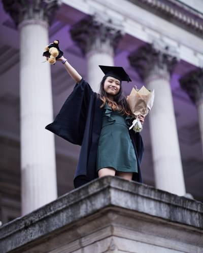 A low angle photo of a student in graduation robes and a teal dress stands on a plinth in front of the classical columns of the UCL Portico. She holds a bouquet of flowers and a teddy bear in an outstretched arm above her head.