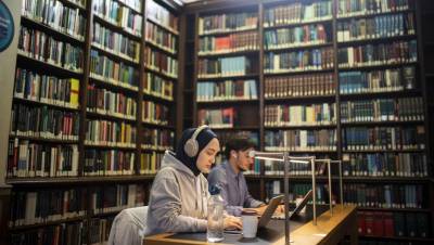Two students are studying on their laptop computers in the book-lined library. One student has a beard and is wearing a blue shirt. The other student is wearing a grey hoodie, a hijab, and large headphones.