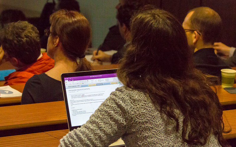 Students sit in a lecture theatre with their backs to the camera. A female student takes notes on her laptop, her screen facing the camera.