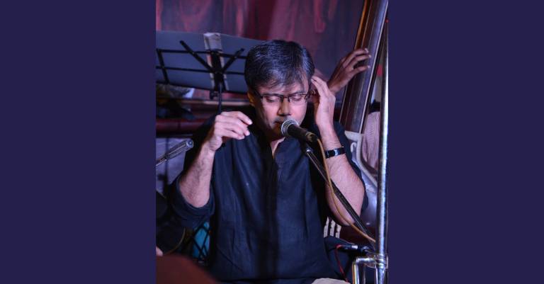 In the image you see Amit Chaudhuri performing, singing into a microphone in front of him, eyes closed. He wears a dark blue shirt and in the background is a music stand 
