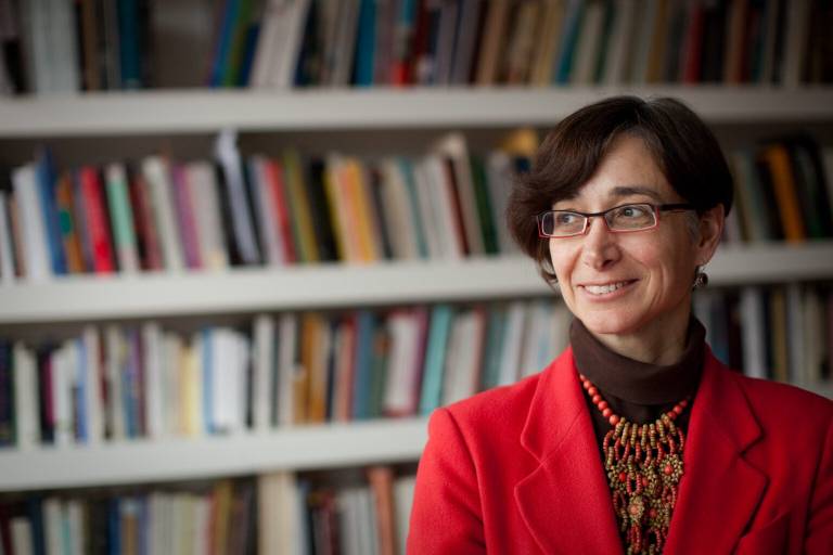 Portrait of Tamar Herzog standing in front of fully stocked bookshelves. She stands in the right half of the image looking to the left. She wears a bright red jacket, dark brown turtleneck, glasses, her hair is dark brown and shortish.