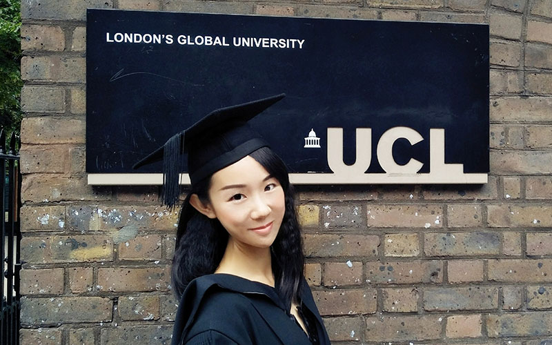 A photo of Luna Zheng in her graduation robes smiling in front of a UCL sign. Permission: Luna Zheng.