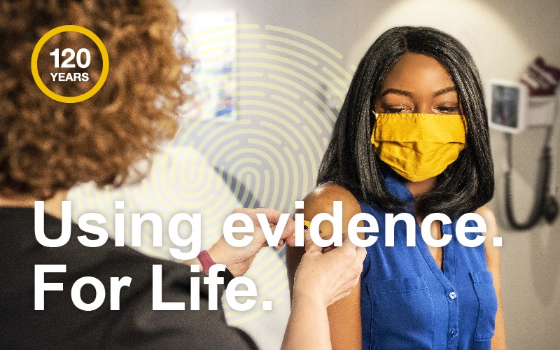 Nurse placing sticker on patient following vaccination with 120 roundel and white text 'Using evidence. For Life'