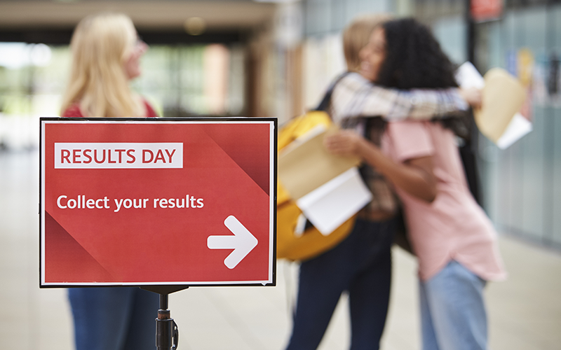 Exam results day red collection sign and blurred figures of students hugging. Credit: Monkey Business / Adobe Stock Standard License.