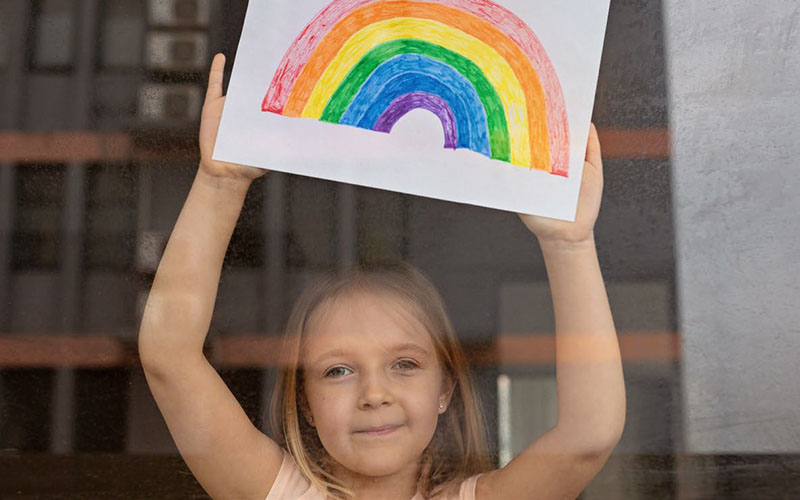 A girl holding up a hand drawn rainbow up against a window