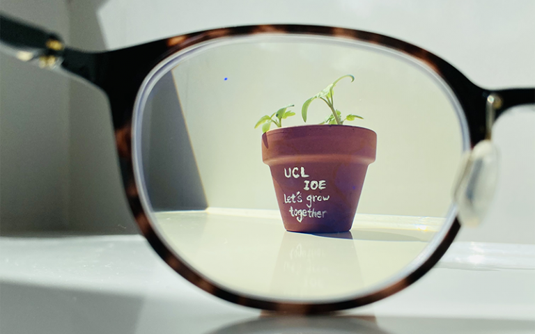 A brown clay flower pot with a green seedling and the words 'UCL IOE: let's grow togther' written in white, viewed through the lens of a pair of glasses laid on a white desk.