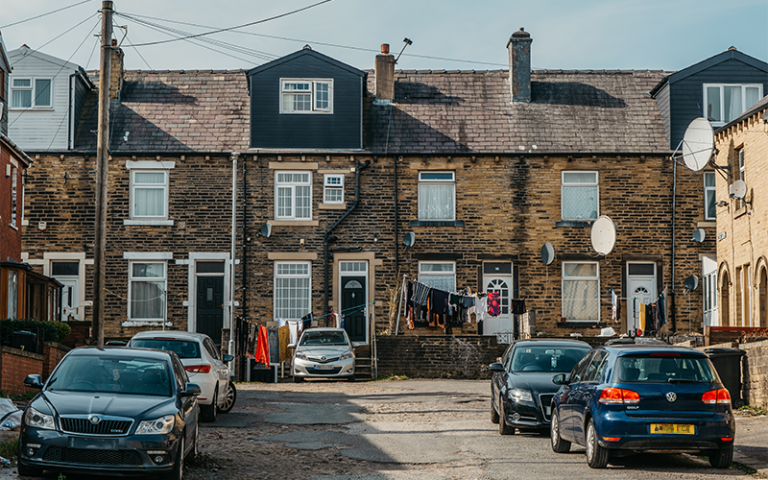 Narrow two and three story terraced houses of Halifax's Park Ward with laundry hanging outside, with cars parked on the sides of a roughly tarmacked road