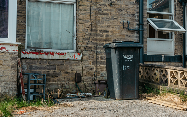 A black refuse bin stands outside a brick house in poor repair with weeds and peeling paintwork.