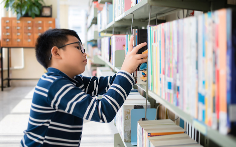 Boy choosing a fiction book from school library shelves