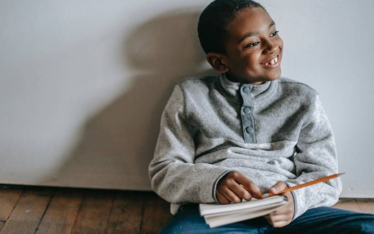 Child smiling and sitting against a wall with notebook in hand. Image: Katerina Holmes via Pexels