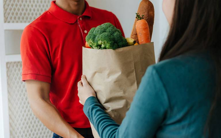 Food delivery service man in red uniform handing fresh food to young woman. Photo: Vittaya_25 / Adobe Stock