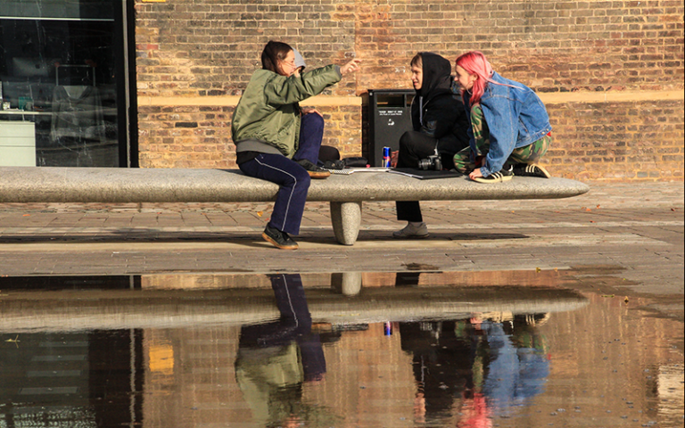 Three young adults on a stone bench reflected in a puddle at Granary Square, Kings Cross.