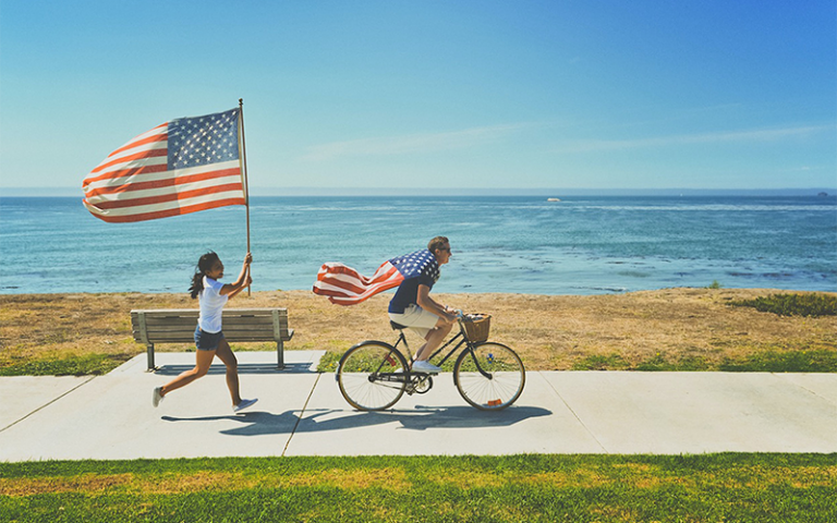 Woman running and man cycling along a waterfront, each holding a USA flag