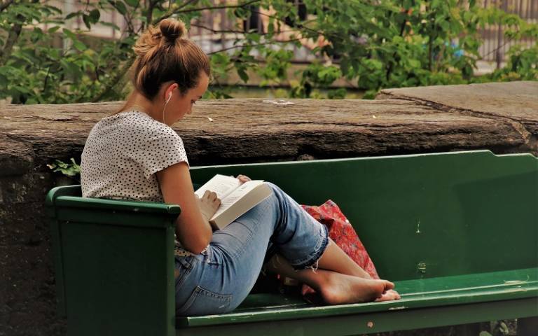 Female student reading outside on a bench with headphones in. Image by pasja1000 from Pixabay.