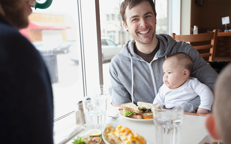 Baby sitting on happy father's lap at lunch. Hero Images via Adobe Stock