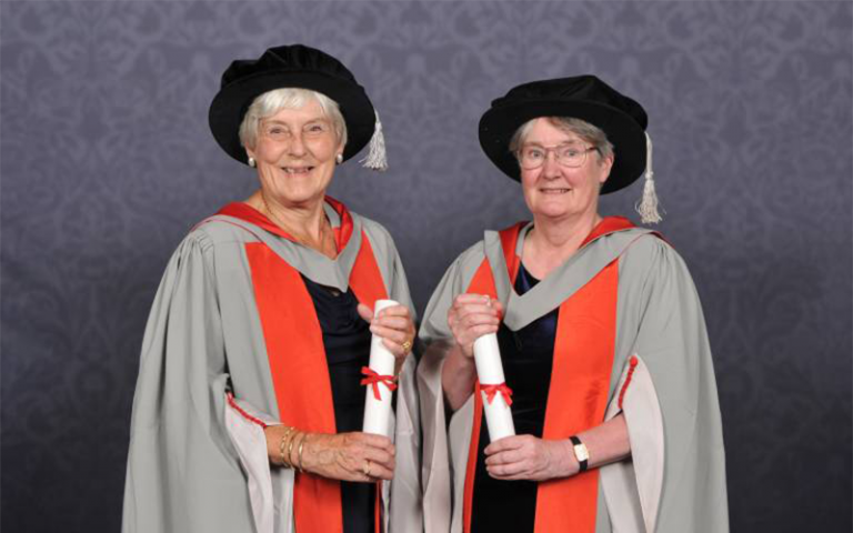 Two women wearing doctoral graduation robes holding diplomas. Photo by Tempest for UCL