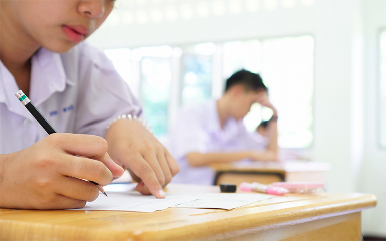 GCSE pupils wearing lilac school uniform shirts fill out test papers at wooden desks