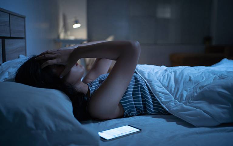 Young person lies in bed covering their face with a mobile phone beside them (Photo: torwaiphoto / Adobe Stock)