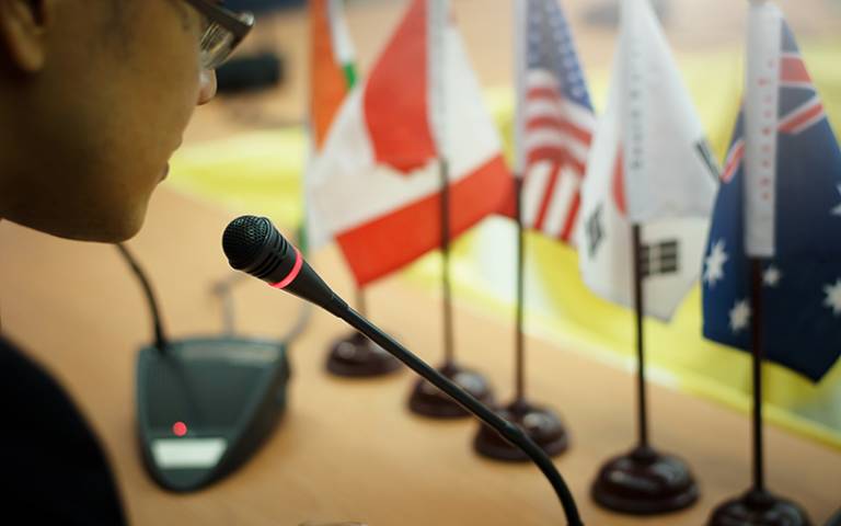 A man wearing glasses speaks into a microphone with a number of country flags in front of him. (Photo: khampiranon / Adobe Stock)