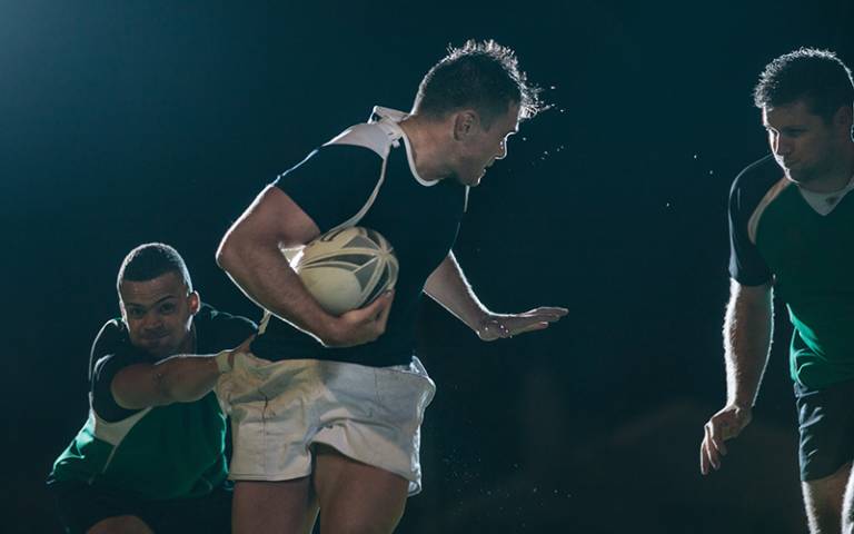 Three men playing rugby. Man holding the ball is holding up his left arm to fend off an opponent whilst grabbed around the waist by another player. (Image: Jacob Lund / Adobe Stock)