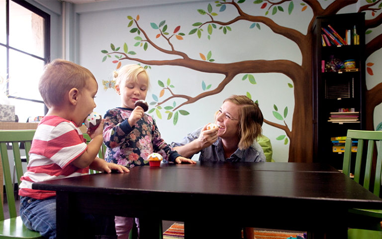 Woman in a nursery classroom laughing as two children pretend to eat toy cupcakes. Credit: Cavan for Adobe.
