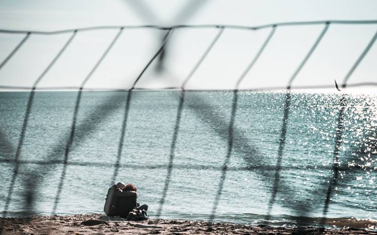 Two figures in the distance sitting on sand with wire fencing in the foreground. (Photo: Edward R / Adobe Stock)