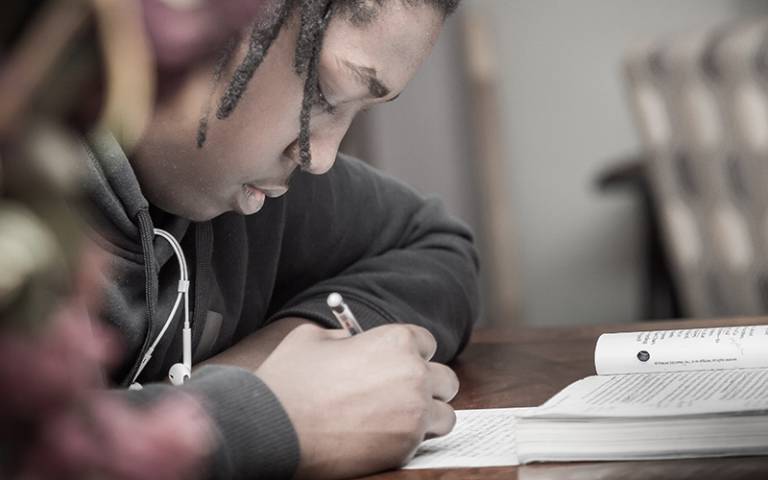Black teenage boy writing at wooden desk.