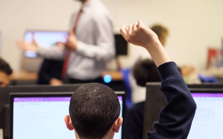 Student raises hand in foreground, teacher in background. Image: Phil Meech for UCL Institute of Education