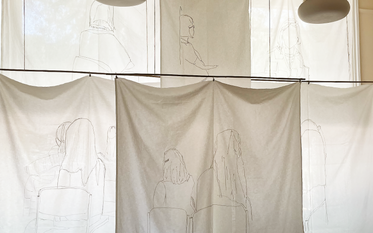 Sails by Nora Wuttke. White cloth hangs like ship's sails. They are embroidered with line drawings of people. Image permission: Nora Wuttke.