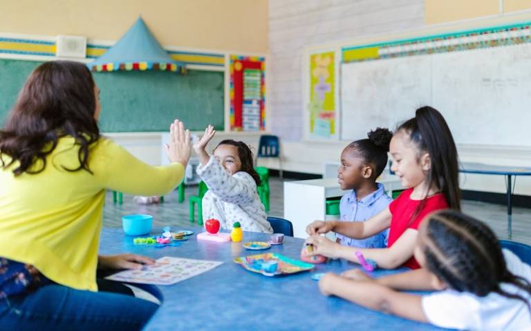 Teacher high fiving child with other nursery pupils sat around table