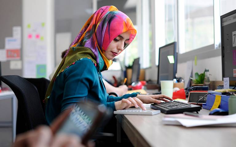 A woman wearing a colourful headscarf is working at a desk in an office. (Photo: Rawpixel.com / Adobe Stock)
