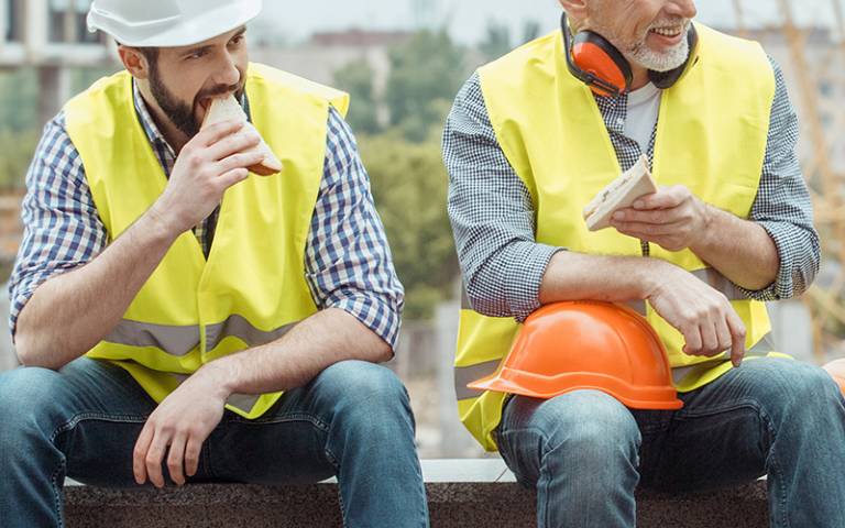 Two men in hi-vis vests and construction workwear eating sandwiches. (Photo by Friends Stock / Adobe Stock)