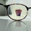A brown clay flower pot with a green seedling and the words 'UCL IOE: let's grow togther' written in white, viewed through the lens of a pair of glasses laid on a white desk.