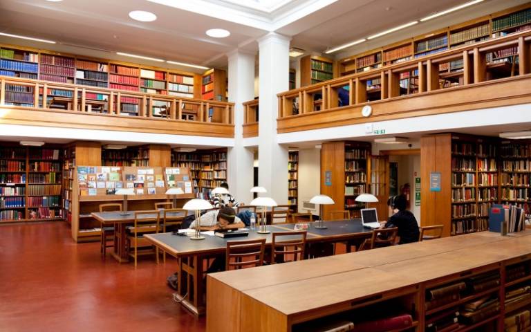 Three students are sitting at different tables in the main UCL library. There are wooden bookshelves around them