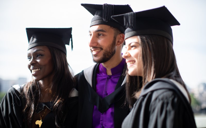 A group of three students wearing graduation caps and gowns looking to the left and posing for a photo