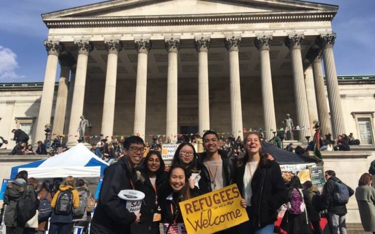 A group of students posing outside the UCL Portico with a yellow sign saying 'Refugees welcome'
