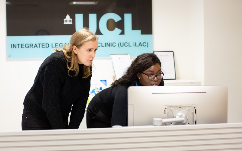 Two members of staff at the UCL Integrated Legal Advice Clinic, looking at a computer at the reception desk. Behind them is a sign on the wall with the UCL logo