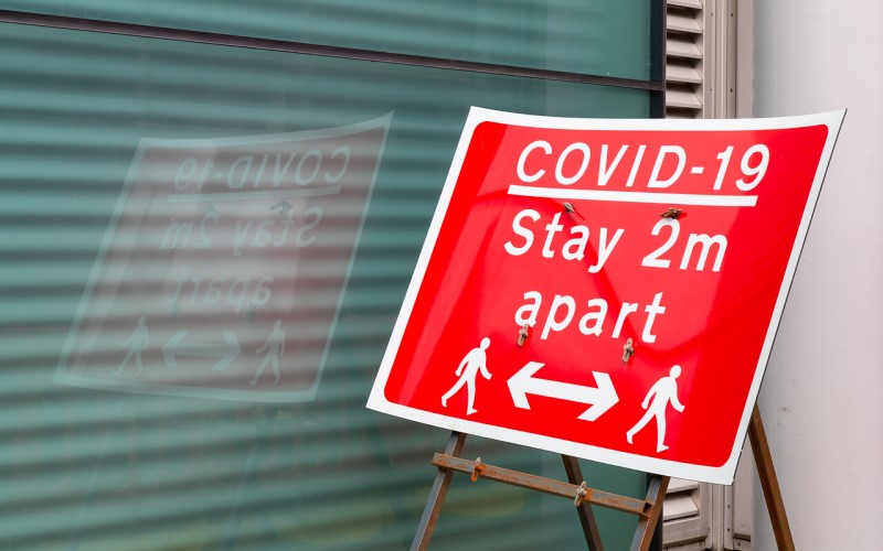 A red sign with white text: "Covid 19 - stay 2m apart", with symbols of two people separated by a horizontal arrow